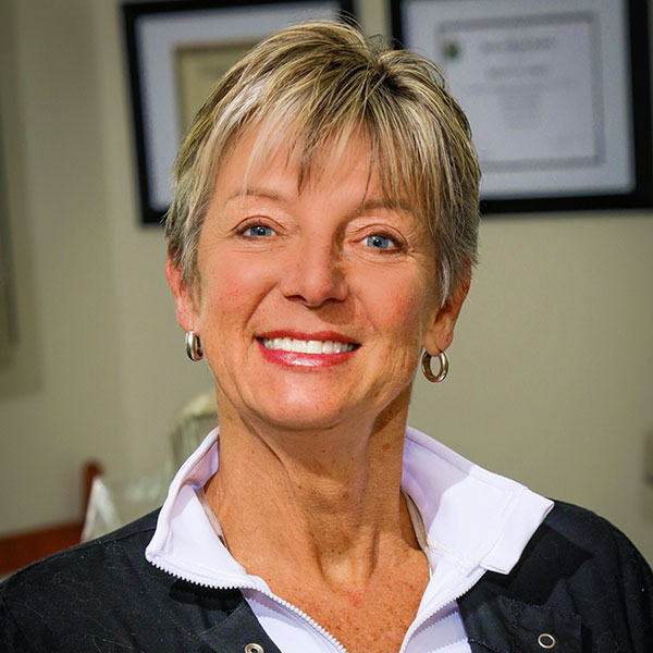 Smiling woman with short blonde hair wearing a white collared shirt and black vest, indoor office background with certificates on the wall