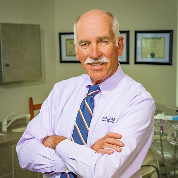 Older man with a white mustache in a lavender dress shirt and striped tie, arms crossed in a medical office with certificates on the wall.