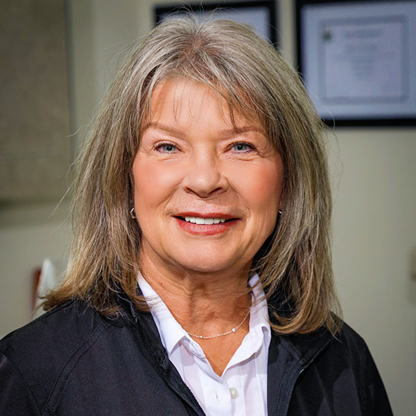 Smiling middle-aged woman with shoulder-length gray hair, white blouse and black jacket, office background with framed certificates.