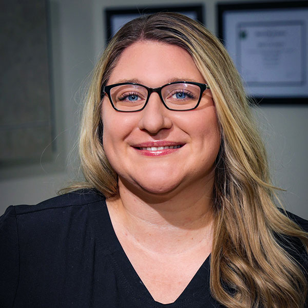 Smiling woman with blonde hair and black-framed glasses, wearing a dark blouse in an office setting.