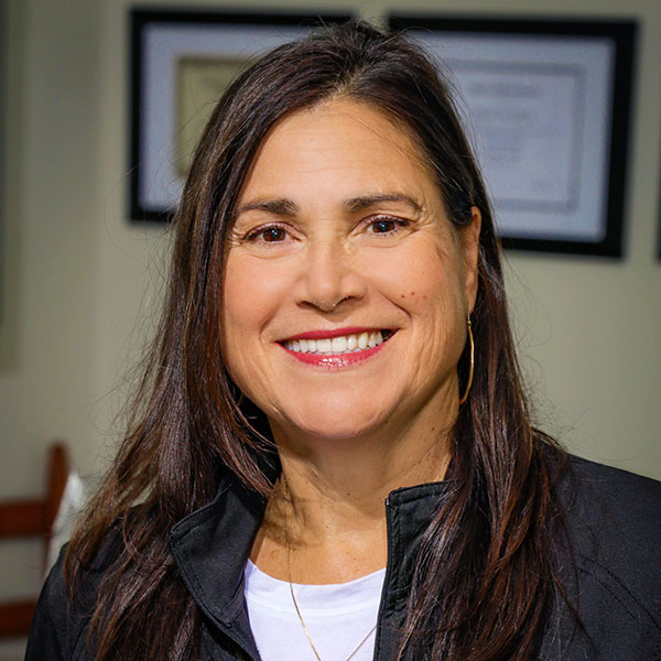 Smiling woman with long dark hair wearing a white top and black jacket, indoors with framed certificates in the background.