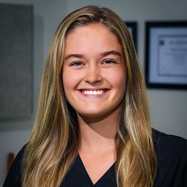Smiling young woman with long blonde hair in a black top, office background behind her.